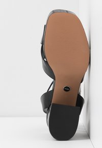 Vue unique d'une sandale à talon noir avec semelle texturée marron clair et talon bloc reposant contre un fond blanc en coin.