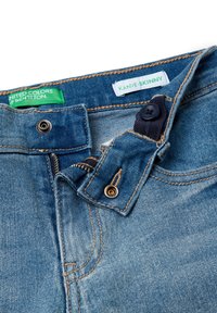 Jeans de mezclilla azul con cremallera parcialmente abierta, cierres de botón y presión, y etiquetas de marca en verde y blanco en el interior de la cintura.