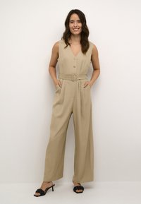 Salopette beige con scollatura a V, chiusura con bottoni e gambe ampie. Presenta una cintura in vita e tasche laterali. Indossata con sandali neri con tacco.