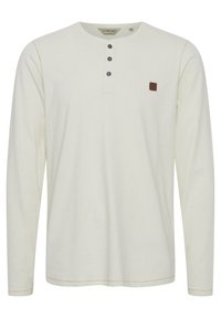 Camisa henley de manga larga en un tono beige claro, con una tapeta de tres botones y un pequeño parche de logo marrón en el lado izquierdo del pecho. Textura suave, diseño casual.