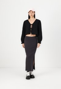 Blusa nera cropped con laccetto davanti, maniche lunghe a sbuffo. Gonna maxi grigia scura con spacco laterale. Calze bianche e scarpe platform nere. Cappellino rosa.