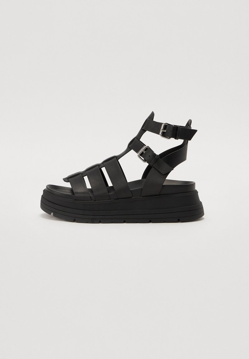 Musta korkea sandaalit, jotka on valmistettu synteettisestä materiaalista useilla hihnoilla ja säädettävillä soljilla. Paksu pohja, jossa on teksturoitu kuosi.