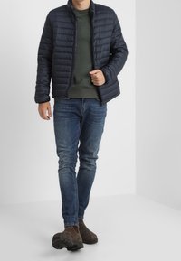 Marinblå pufferjacka med ståkrage, horisontellt kviltad design, bärs över en mörkgrön tröja, matchad med blå jeans och bruna ankelstövlar.