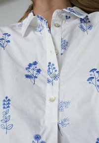 Hvid skjorte med knapper, der har blå blomsterbroderi, klassisk krave og perleknapper. Glat bomuldsstof med detaljerede plante Designs.