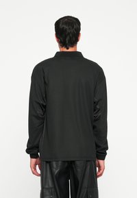 Schwarzes Langarmshirt mit Polokragen, aus glattem Stoff und mit strukturierten Akzenten an den Ärmeln. Kombiniert mit schwarzen Lederhosen.