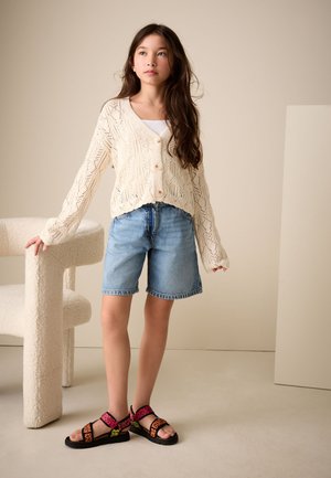 Fiatal lány hosszú barna hajjal, krémszínű kötött kardigánt, farmer sortot és színes, leopárdmintás szandált visel, egy textúrált fehér szék mellett áll.