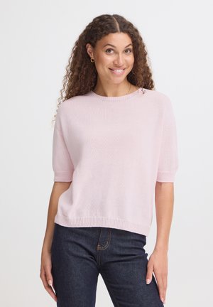 Kvinde med krøllet hår iført en lyserød kortærmet sweater og mørkeblå jeans, smilende og poserer mod en hvid baggrund.
