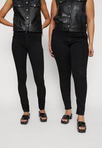 Coletes curtos de couro preto com bolsos frontais, combinados com jeans skinny pretos e sandálias de salto bloco pretas. Textura suave por todo o conjunto.