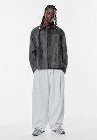 Grå jakke lavet af blødt materiale med en frontlomme, parret med løstsiddende, lysegrå sweatpants og grå sneakers.