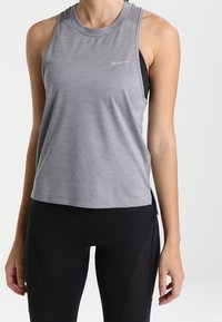 Graues Sporttop mit rundem Ausschnitt, mit kleinem Logo und seitlichen Schlitzen, kombiniert mit schwarzen Leggings mit Mesh-Details.