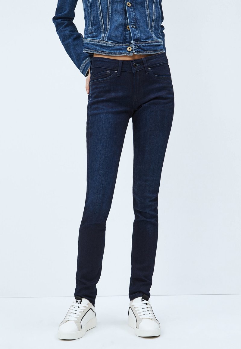 Dunkelblaue Skinny-Jeans mit Fronttaschen, minimaler Nahtführung und glatter Textur, kombiniert mit einer Jeansjacke und weißen Sneakers.