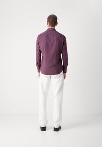 Paarse langmouwshirt, met knoopmanchetten, op maat gesneden pasvorm. Gecombineerd met witte broeken en zwarte schoenen. Minimalistisch design, gladde textuur.