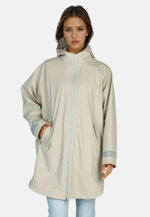 Woman wearing light beige hooded raincoat with front zipper and side pockets, standing against plain background.