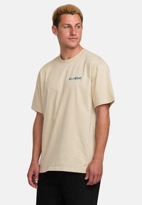 Beige cotton t-shirt with a relaxed fit, short sleeves, and a small green logo on the left chest. Smooth texture, simple design.