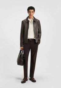Veste en cuir marron, chemise à zip crème, pantalon sombre et chaussures marron. Le mannequin tient un sac sombre, se tenant devant un fond neutre.