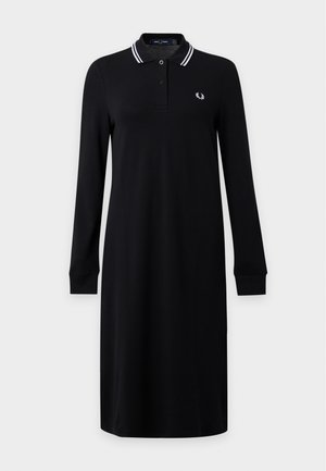Femme portant une robe polo noire à manches longues avec un col rayé blanc, des chaussettes blanches et des mocassins noirs, debout les mains dans le dos.