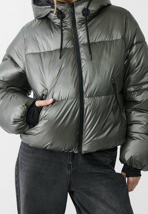 Doudoune grise brillante à capuche avec fermeture éclair noire et cordons réglables, portée avec un jean noir, main dans la poche de la veste.