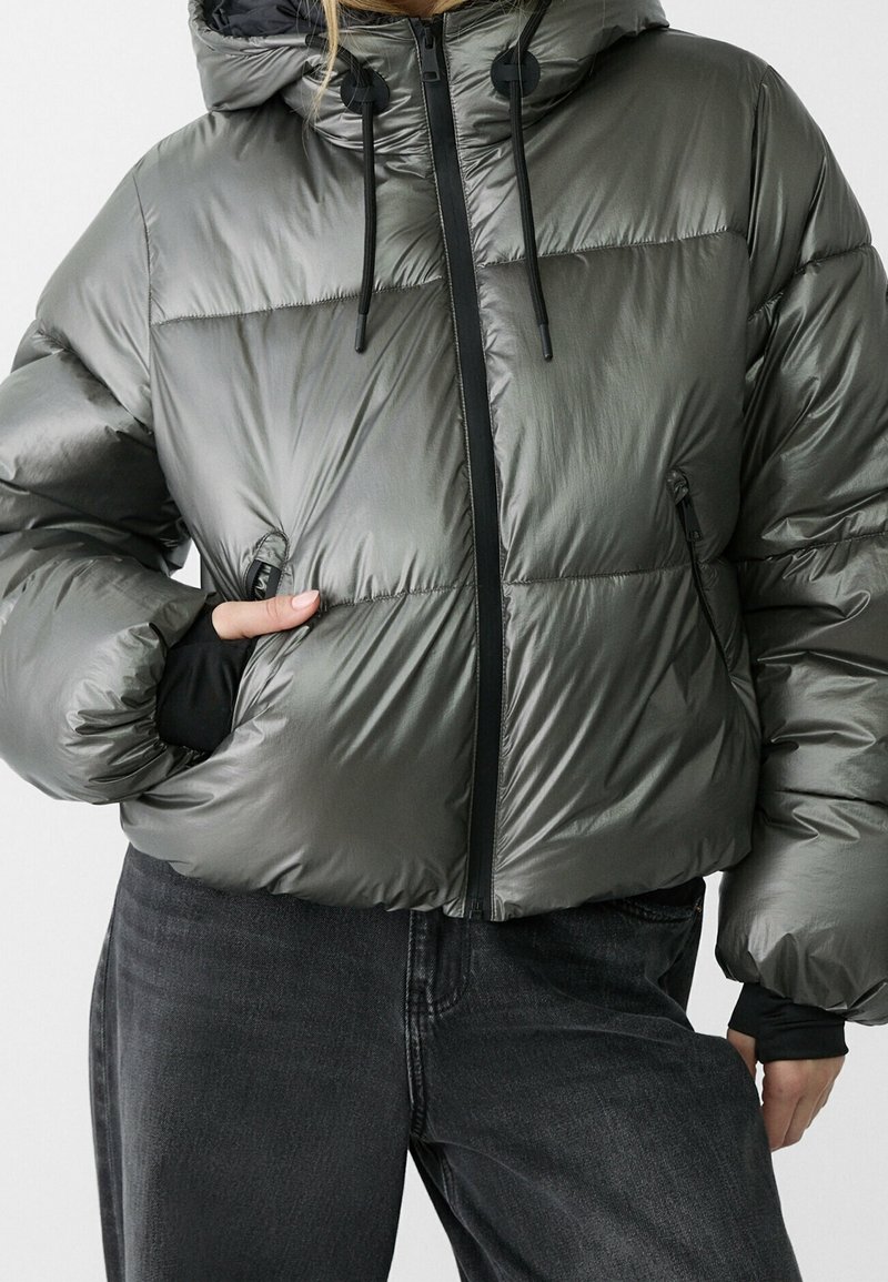 Doudoune grise brillante à capuche avec fermeture éclair noire et cordons réglables, portée avec un jean noir, main dans la poche de la veste.