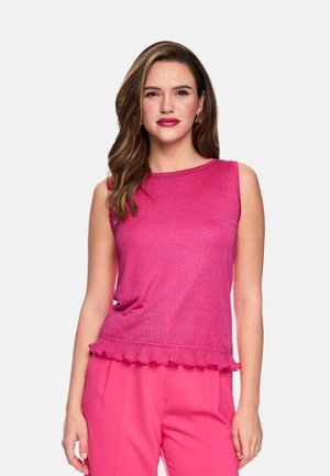 Frau mit langen braunen Haaren trägt ärmelloses pinkes Oberteil mit Rüschen am Saum und passende pinke Hose, stehend vor einem schlichten, hellen Hintergrund.