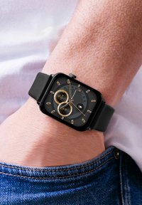 Montre intelligente noire avec un cadran carré, des accents dorés, deux sous-cadrans et un fond noir texturé. Bracelet en caoutchouc, portée au poignet.
