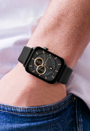 Montre intelligente noire avec un cadran carré, des accents dorés, deux sous-cadrans et un fond noir texturé. Bracelet en caoutchouc, portée au poignet.