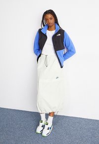 Femeie stând împotriva unui perete alb, purtând o jachetă din polar albastră și neagră, un top alb, o fustă Nike deschisă, șosete albe și teneși Nike albi.