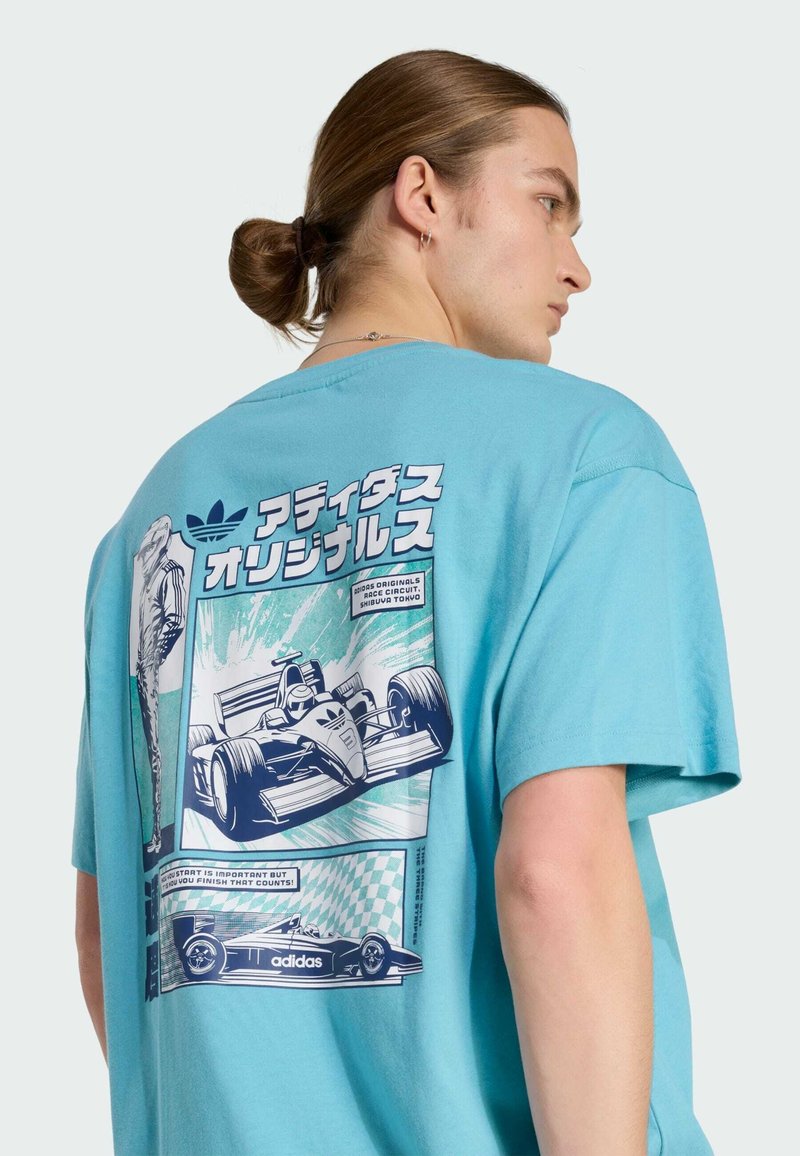 Giovane con i capelli raccolti che indossa una maglietta blu con testo giapponese e illustrazioni dettagliate di auto da corsa sulla schiena.