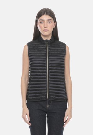 Donna che indossa un gilet trapuntato nero senza maniche, con la cerniera chiusa, in piedi con le braccia rilassate davanti a uno sfondo chiaro e semplice.