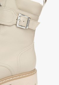 Bottine en cuir beige avec une finition texturée, dotée d'une accent en boucle argentée et d'une semelle épaisse de couleur claire.