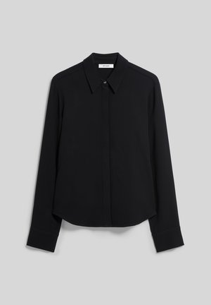 Femme debout devant un fond uni, portant une chemise noire à manches longues, un pantalon noir à jambes larges et des chaussures pointues cloutées.