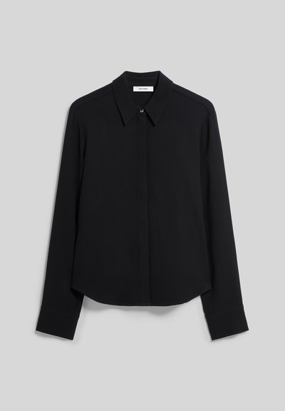 Chemise noire à manches longues avec col, boutons dissimulés et poignets droits, présentée sur un fond gris clair uni.