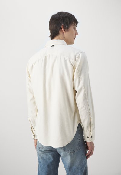 Camisa de manga larga de color crema, hecha de algodón, con un cuello abotonado y un dobladillo redondeado. La parte trasera presenta una costura central y una etiqueta.