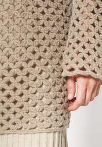 Main avec des ongles naturels à côté d'un pull en laine beige épais au crochet avec un motif floral ajouré sur un tissu côtelé.