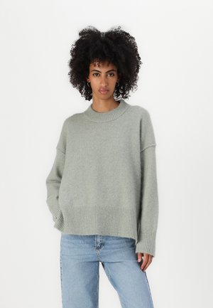 Lys grå genser laget av mykt materiale, med ribbestrikket kant og mansjetter, i en avslappet passform og rund halslinning. Kombinert med blå jeans.