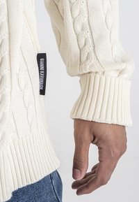 Cremfarbener Zopfmuster-Pullover mit strukturierten Mustern und gerippten Bündchen, mit einem schwarzen Etikett am Ärmel. Getragen mit blauen Jeans.