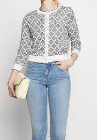 Cárdigan blanco con patrón floral negro, escote redondo y cierre de botones, combinado con jeans azul claro y un pequeño bolso amarillo.