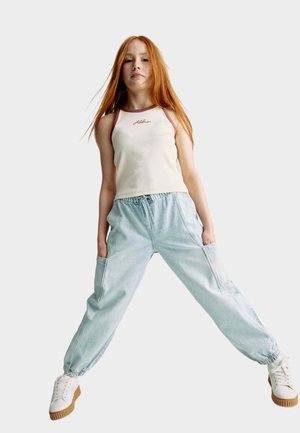 Licht crèmekleurig tanktop met bruine biezen en geborduurd logo, gecombineerd met lichtblauwe, loszittende broek met zijzakken. Witte sneakers.