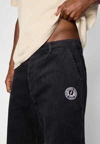 Pantalon en velours côtelé noir avec des rayures verticales, arborant un écusson rond blanc sur la cuisse gauche et des poches standard. Porté avec une chemise beige.