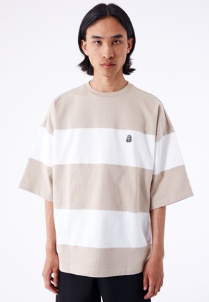 T-shirt surdimensionné beige et blanc rayé avec un col rond et des manches courtes ; arbore un petit logo noir sur la poitrine gauche.