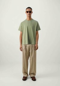 Ανοιχτό πράσινο βαμβακερό t-shirt με στρογγυλή λαιμόκοψη και μικρό λογότυπο, συνδυασμένο με μπεζ χαλαρές παντελόνες και καφέ δερμάτινα παπούτσια.