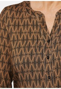 Brauner Stoff mit einem schwarzen geometrischen Baum-Muster, mit einem runden Kragen und Knopfdetails. Weiche Textur, taillierte Passform.