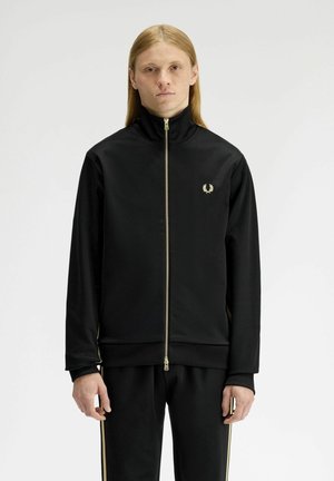 Giovane con capelli lunghi che indossa una giacca nera con zip e pantaloni abbinati con strisce dorate, in piedi contro uno sfondo bianco uniforme.