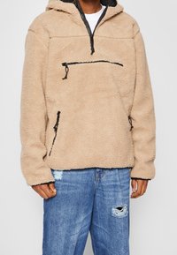 Pullover in felpa beige testurizzata con cappuccio, dotato di una zip frontale nera a metà e due tasche laterali con zip. Indossato con jeans strappati blu.
