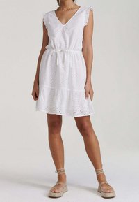 Weißes Kleid mit Lochstickereien, V-Ausschnitt, Rüschenärmeln und gestuftem Saum; tailliert mit Kordelzug. Getragen mit beigen Sandalen.