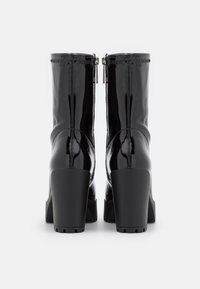 Une paire de bottines noires brillantes avec des talons blocs épais et des zips arrière, présentée de l'arrière sur un fond blanc.