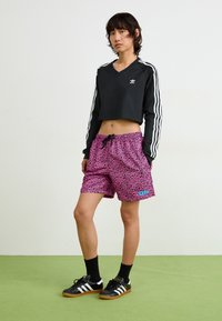 Top preto cropped de mangas longas com três riscas brancas, combinado com calções rosa com padrão de leopardo e um logo azul, e sapatilhas pretas.