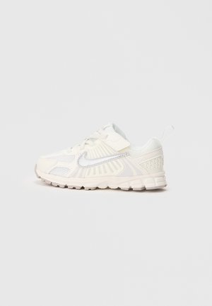Bijele i bež Nike atletske tenisice s mrežastim panelima, vezicama, Velcro trakama i petljom na jednostavnoj svijetloj pozadini.