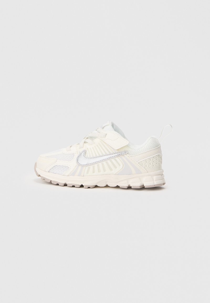 Balti ir kreminiai Nike sportiniai batai su tinklelio panelėmis, raišteliais, Velcro dirželiu ir traukimo juostele ant vientiso šviesaus fono.