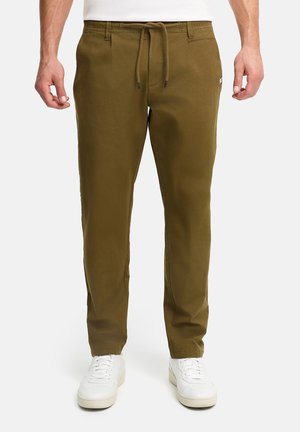 Mann iført olivengrønne bukser med snor og hvite joggesko, stående med armene avslappet ned langs sidene.