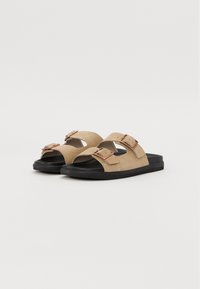Beige semsket sklie sandaler med to justerbare spenner og en svart gummisåle. Har åpen tå-design og flat konstruksjon.